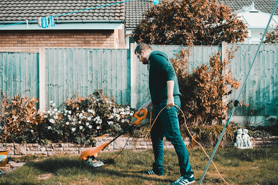 A man wearing glasses, a dark hoodie, and jeans is operating a petrol-powered grass trimmer in a well-maintained garden. The garden features a lush green lawn with areas of freshly cut grass, bordered