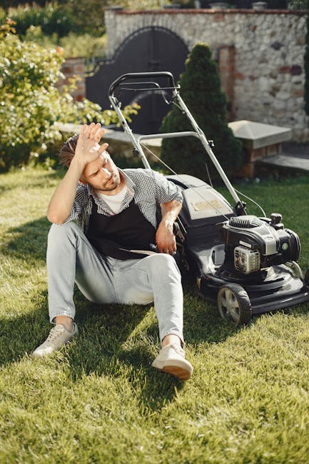 A man wearing glasses, a dark hoodie, and jeans is operating a petrol-powered grass trimmer in a well-maintained garden. The garden features a lush green lawn with areas of freshly cut grass, bordered