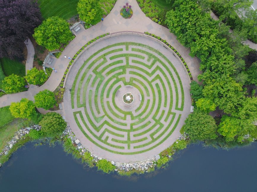 An aerial view of a circular garden featuring a intricate maze with low green hedges and narrow pathways, surrounded by lush green trees. The maze is centered around a small fountain or statue at the