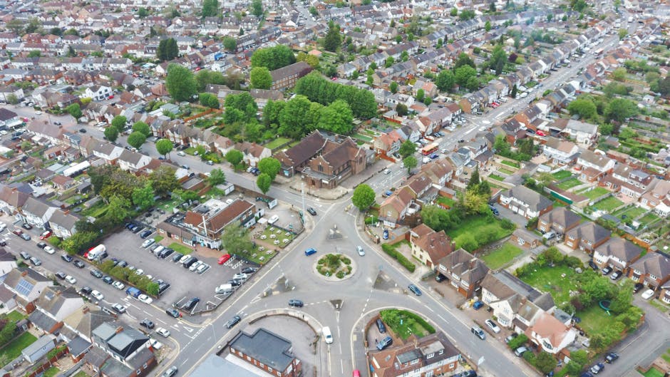 Aerial view of a residential neighborhood featuring a roundabout at the intersection of multiple streets. Surrounding the roundabout are various houses with pitched roofs, small front yards, and garde
