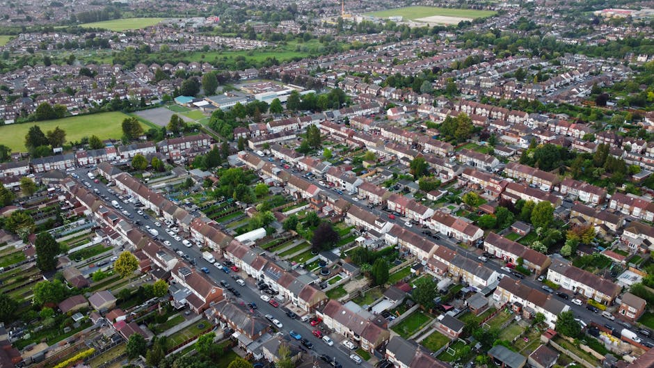 Aerial view of a residential neighborhood featuring a roundabout at the intersection of multiple streets. Surrounding the roundabout are various houses with pitched roofs, small front yards, and garde
